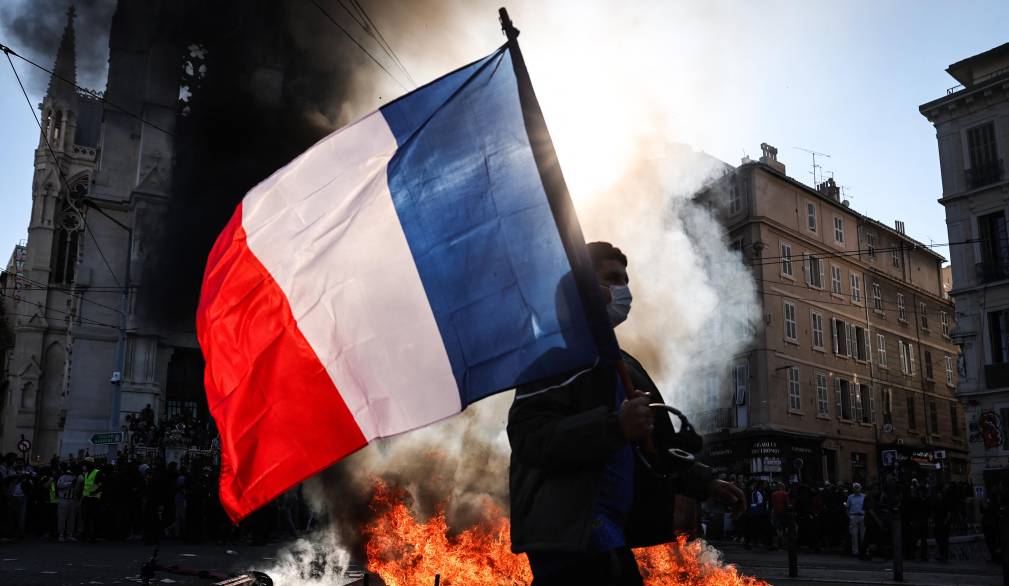 A protester waving a French national flag walks past a fire during clashes prior to the start of a demonstration to protest against the high cost of living, in Marseille, southern France, on October 17, 2026 as part of a nationwide call for mobilisation bringing together various groups expressing their anger: former Yellow Vests, secondary school and university students, climate activists and even farmers. After weeks of student protests, France faces broader, nationwide demonstrations on October 17, 2026 over the cost of living, setting up a potentially fraught six months heading into a presidential election. A police source told AFP that authorities estimated nearly 200,000 people could take part, with 40,000 in Paris. (Photo by Thibaud MORITZ / AFP)